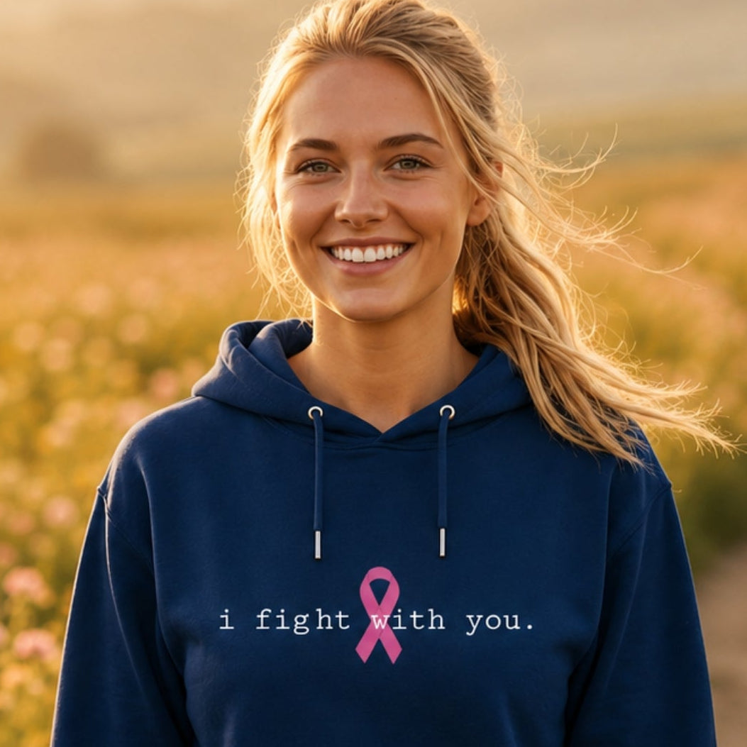 A smiling blonde woman stands in a flower field at sunset wearing a navy blue hoodie with the text 'i fight with you.' and a pink ribbon printed on the chest.