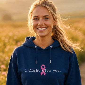 A smiling blonde woman stands in a flower field at sunset wearing a navy blue hoodie with the text 'i fight with you.' and a pink ribbon printed on the chest.