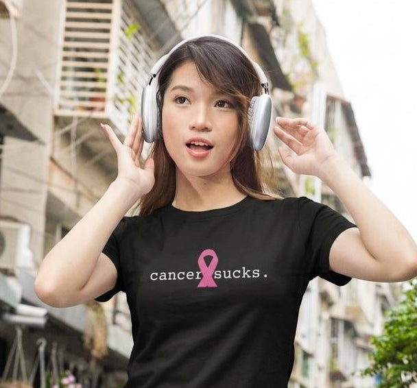 A woman wearing a black t-shirt with a pink ribbon and the text 'cancer sucks.' is shown outdoors with white over-ear headphones.