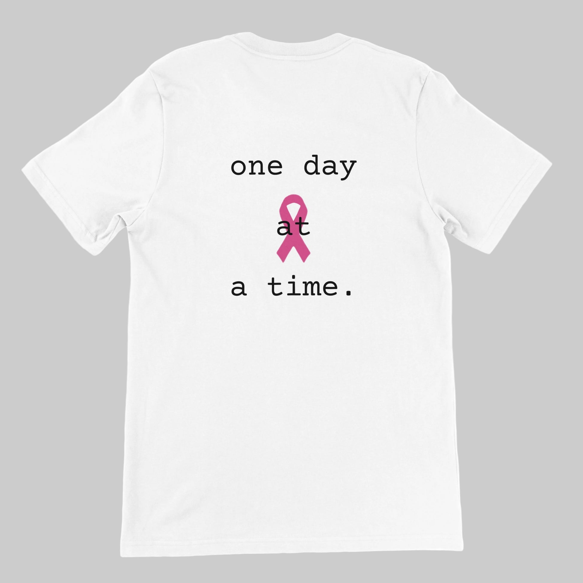 A white short-sleeve t-shirt with a graphic on the front reading 'one day at a time.' in white text, featuring a pink awareness ribbon in the center of the words on a warm grey background