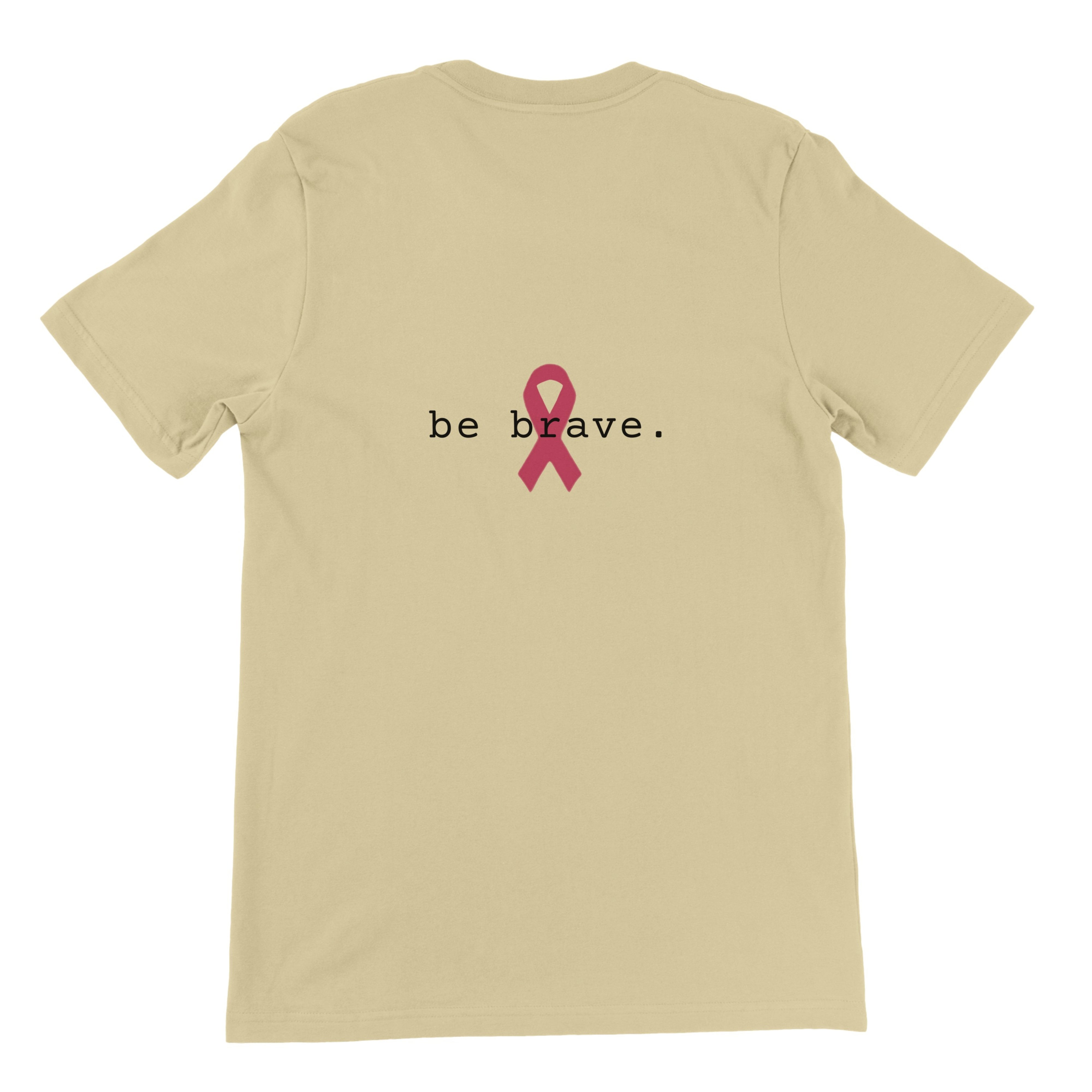 A natural-colored short-sleeve t-shirt is shown from the back, featuring the text 'be brave.' in black with a pink awareness ribbon graphic in the center.