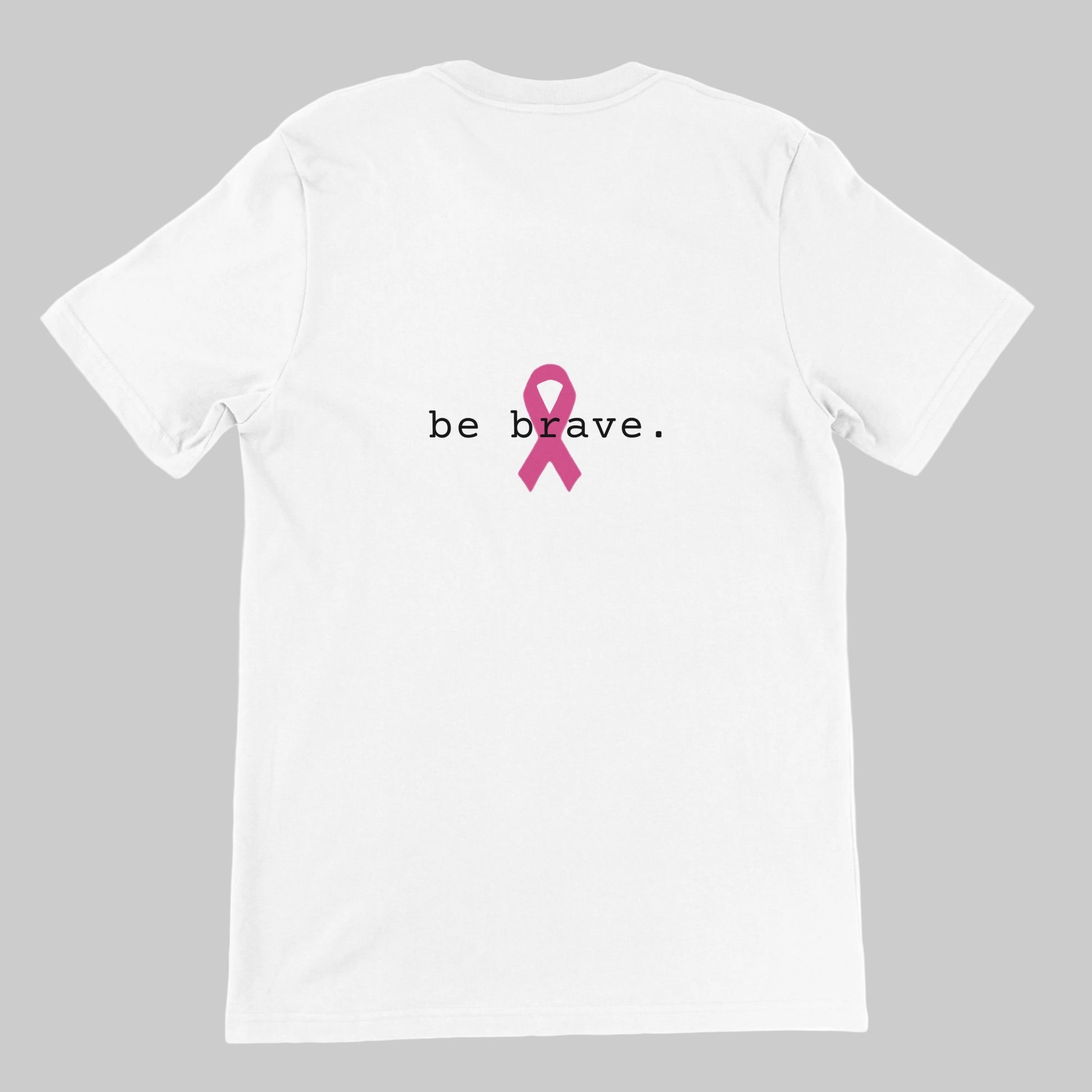 A white short-sleeve t-shirt is shown from the back, featuring the text 'be brave.' in black with a pink awareness ribbon graphic in the center on a warm grey background