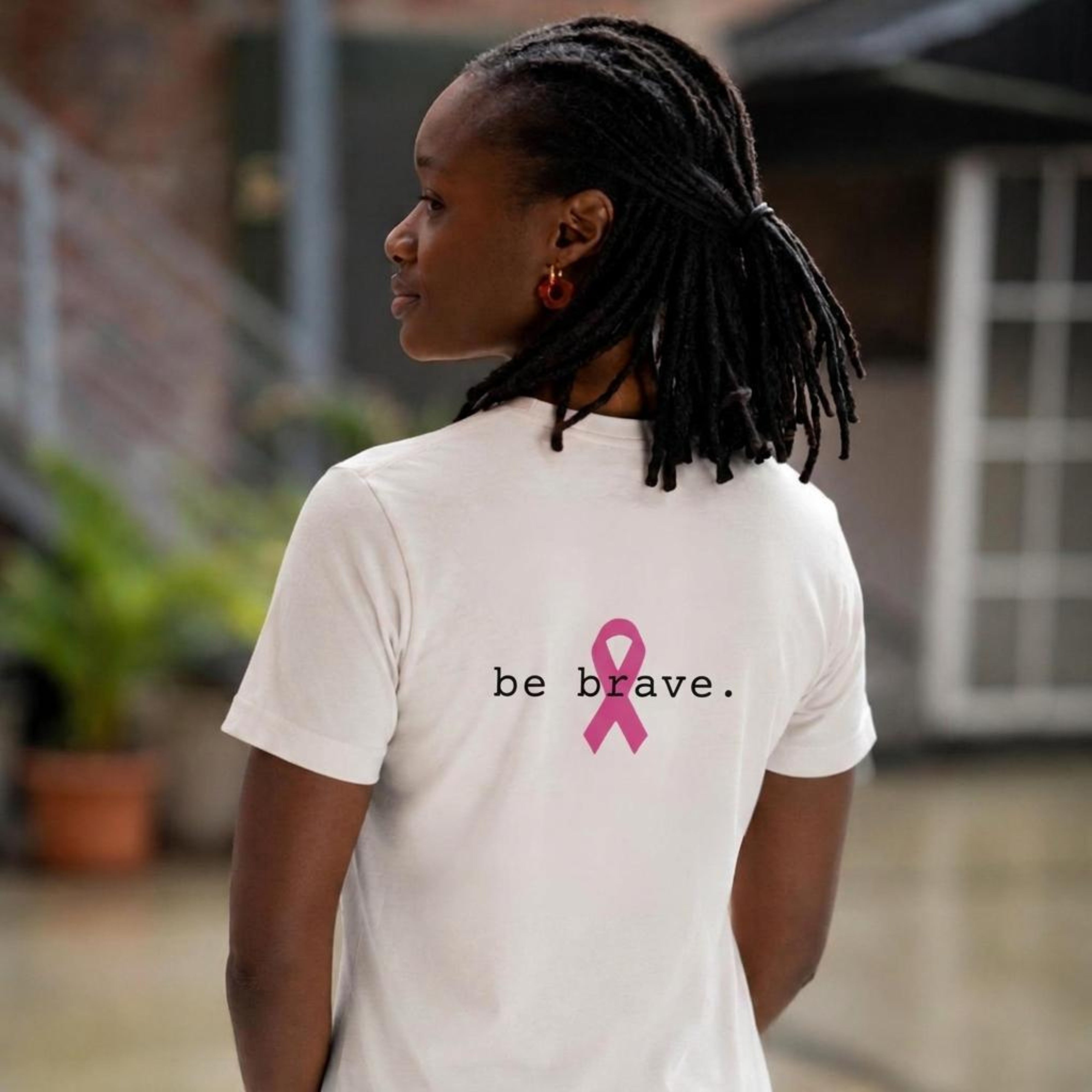 A woman with black dreadlocks is shown from behind wearing a white t-shirt with a pink ribbon graphic and the text 'be brave.' printed on the back, set against a blurred outdoor background.
