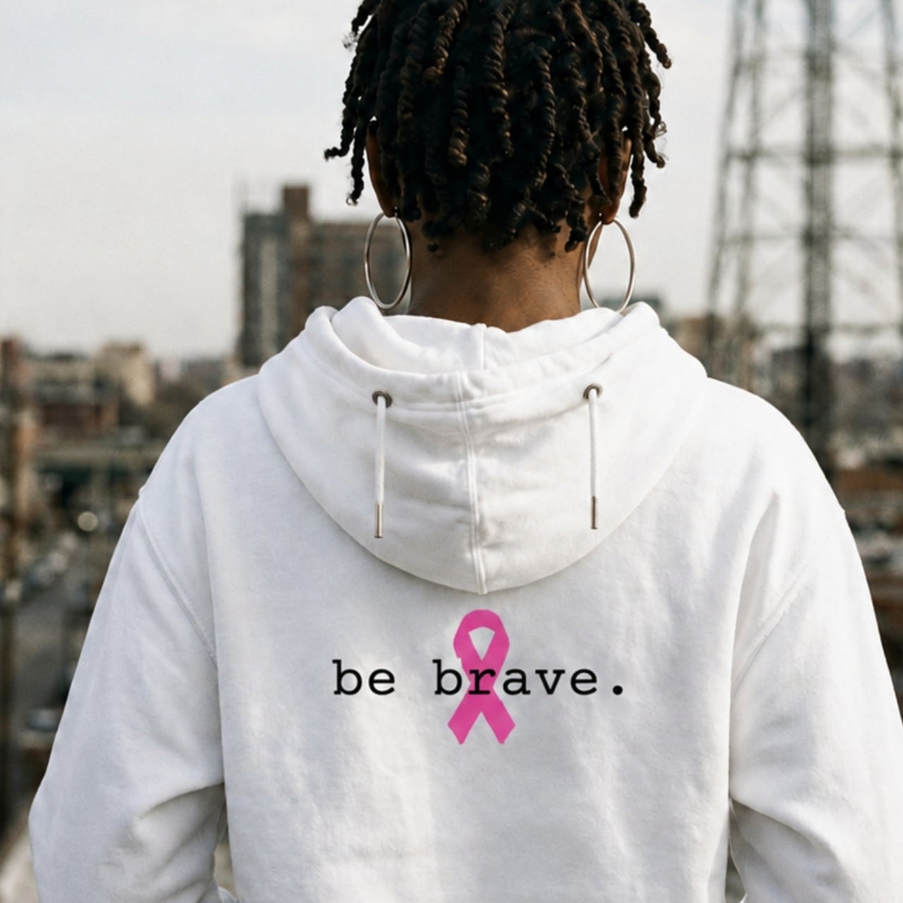 A person viewed from behind wearing a white hoodie with the words 'be brave.' and a pink ribbon printed on the back, standing on a city rooftop with buildings and a tall metal tower in the background.
