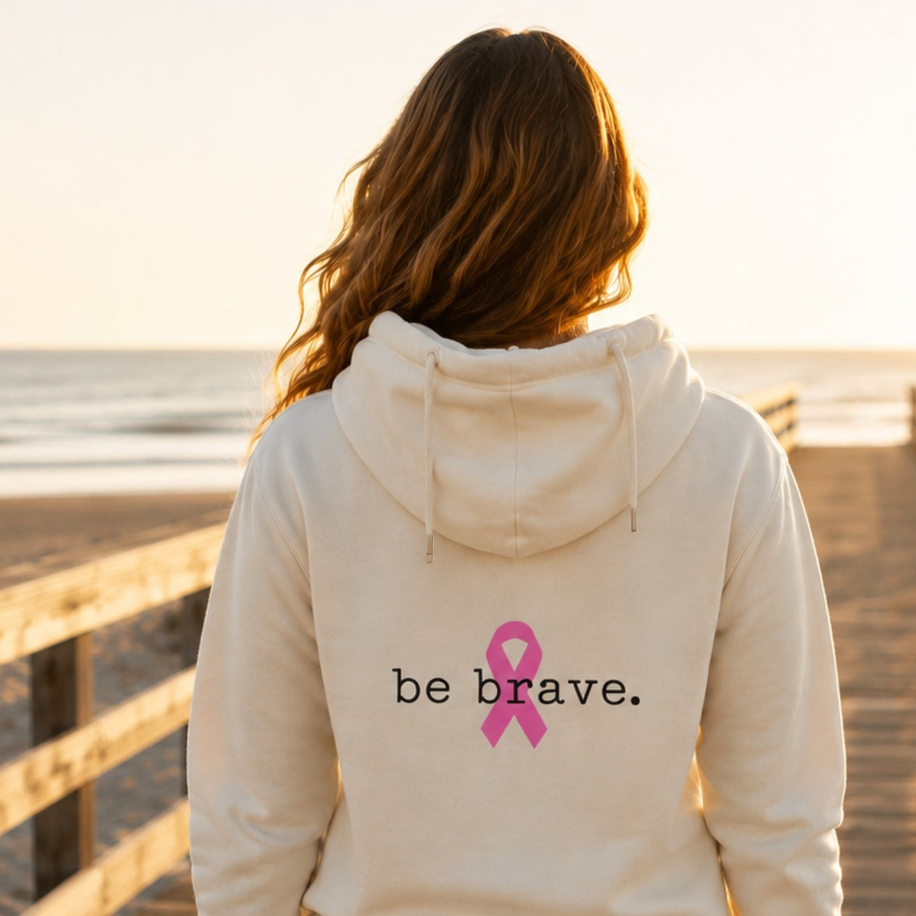 A woman viewed from behind stands on a wooden boardwalk at sunset wearing an bone-colored hoodie with a pink ribbon and the words 'be brave.' printed on the back, paired with blue jeans.