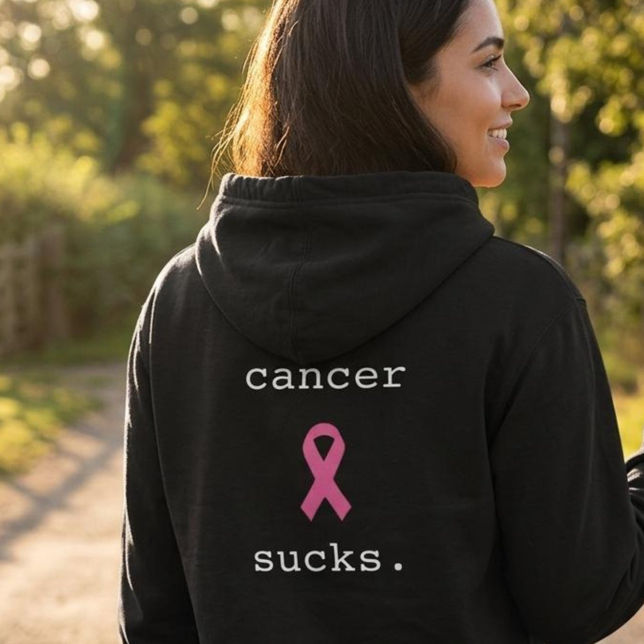 A woman standing outdoors on a sunlit path, shown from the back wearing a black hoodie with white text reading 'cancer sucks.' and a pink ribbon graphic.