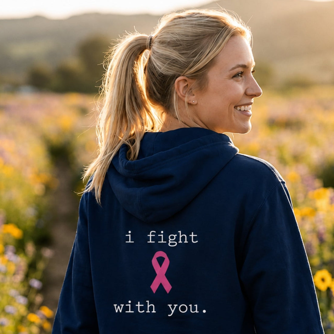 A smiling blonde woman stands in a flower field at sunset wearing a navy blue hoodie with the text 'i fight with you.' and a pink ribbon printed on the chest.