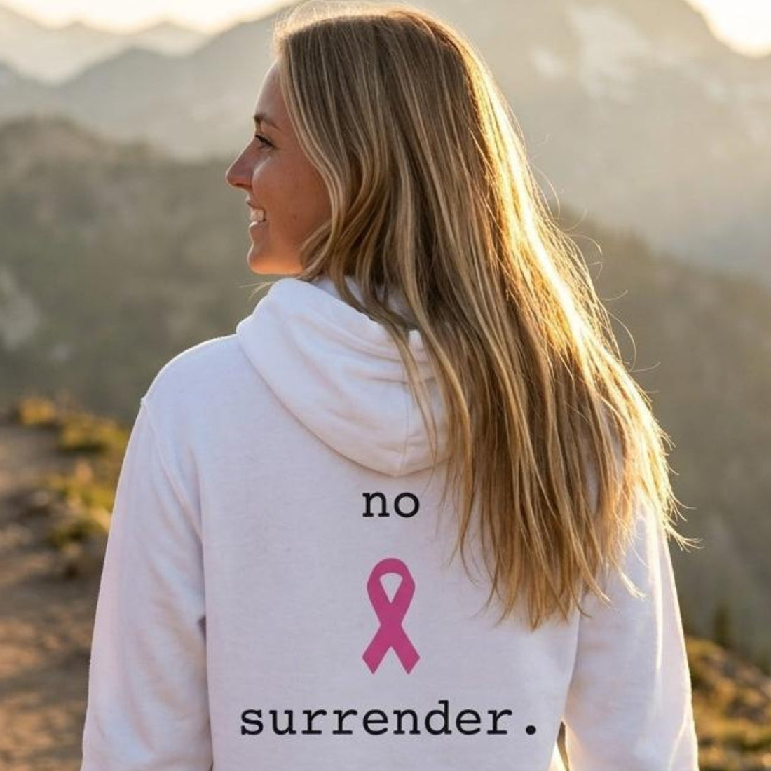 A woman smiling on a mountain trail wearing a white hoodie with the text 'no surrender.' and a pink ribbon graphic on the chest, with pine trees and mountains in the background.