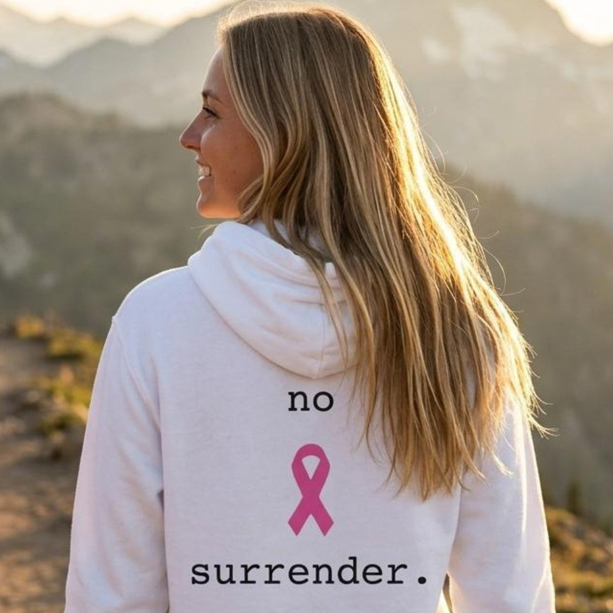 A woman standing on a mountain trail, viewed from behind, wearing a white hoodie with the text 'no surrender.' and a pink ribbon printed on the back.