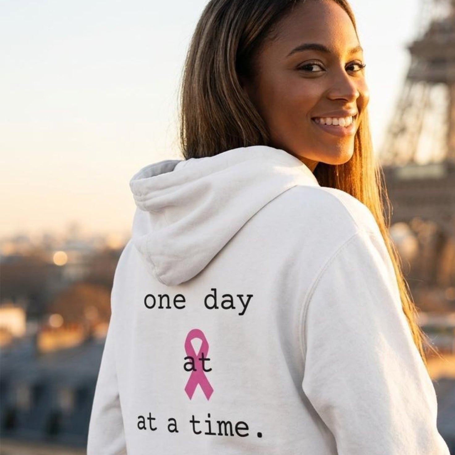 A woman wearing a white hoodie with the text 'one day at at a time.' and a pink ribbon printed on the back, posing with the Eiffel Tower blurred in the background.