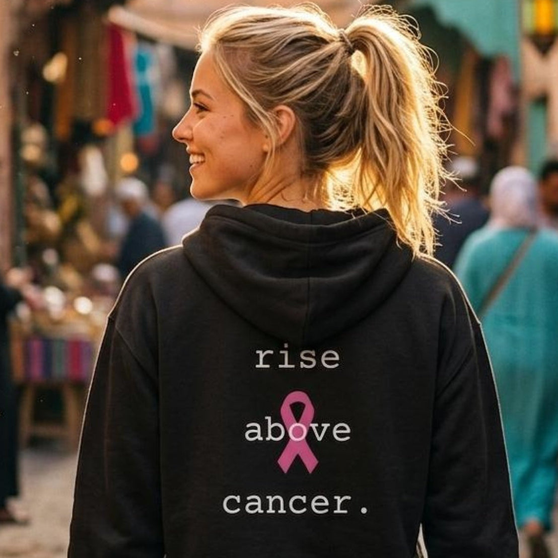 A woman viewed from behind wearing a black hoodie with the text 'rise above cancer.' and a pink ribbon graphic on the back, standing in a sunlit cobblestone street market.