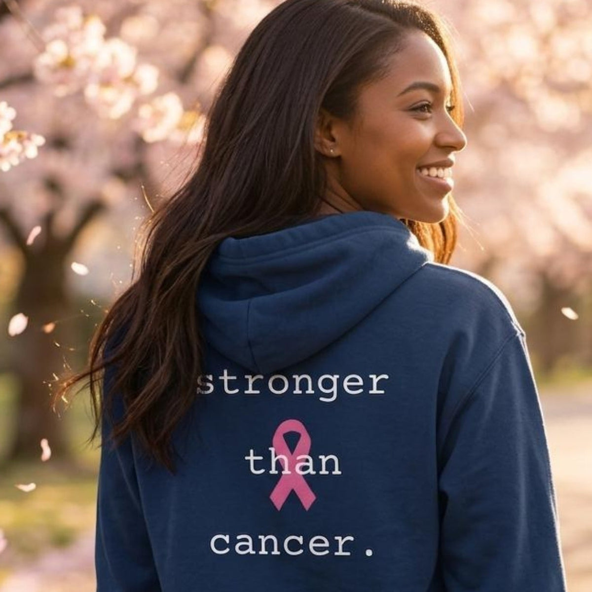 A woman stands outdoors on a path surrounded by blooming cherry blossom trees, wearing a navy blue hoodie from the back. The hoodie features the text 'stronger than cancer.'
