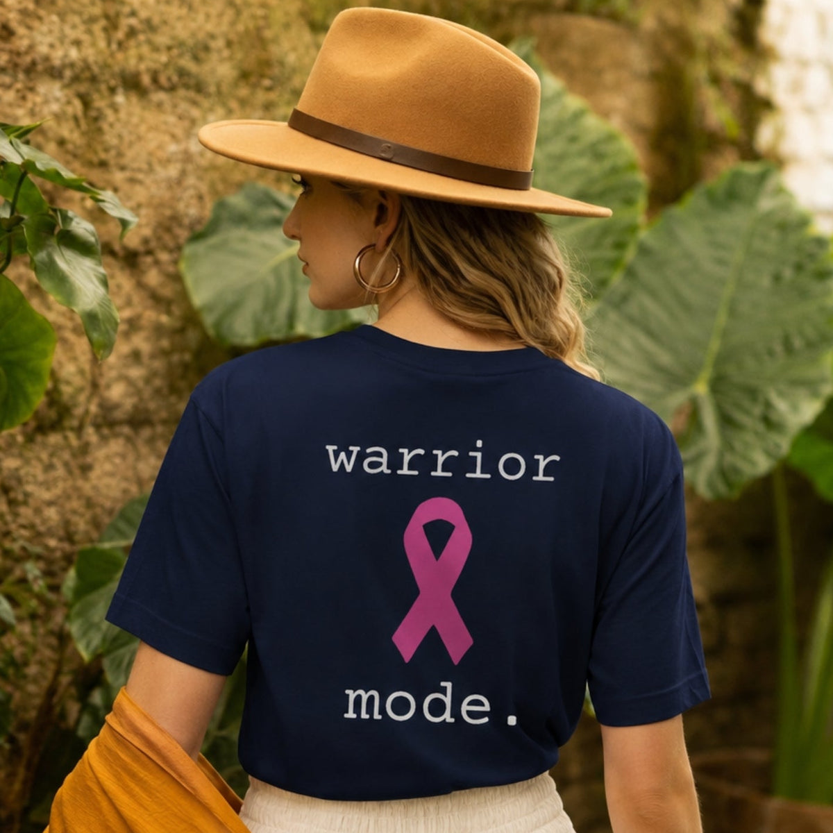 A woman wearing a navy blue t-shirt with a pink ribbon graphic and the words 'warrior mode.' on the back, styled with a tan hat, white pleated skirt, and draped mustard yellow cardigan in a lush garden setting.