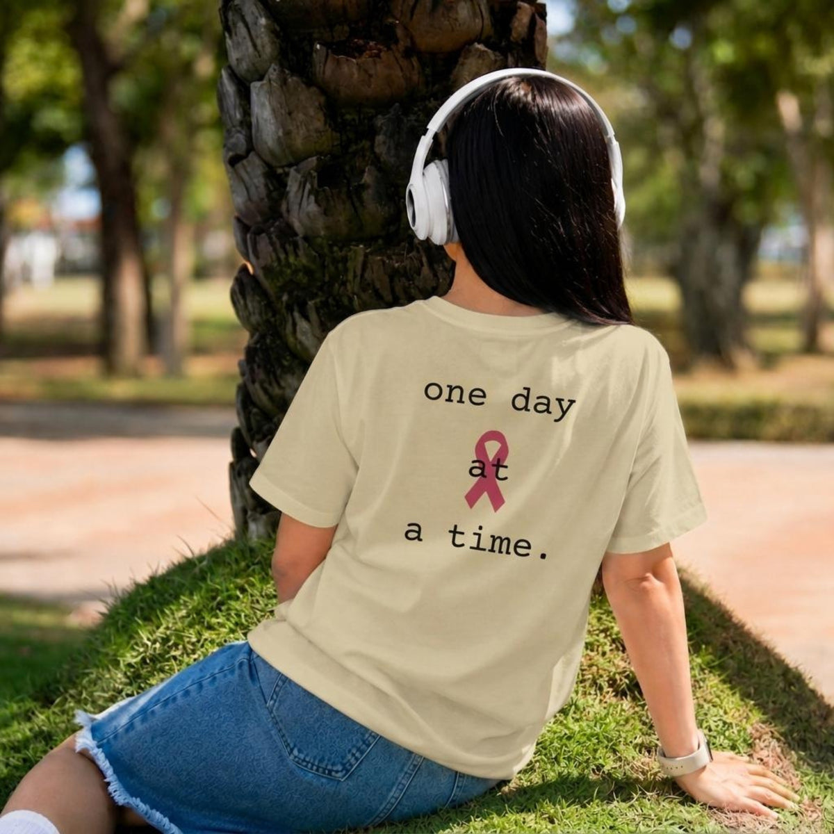 A woman sitting outdoors on grass in front of a tree, seen from behind, wearing a naturalT-shirt with the printed phrase 'one day at a time.' and a pink awareness ribbon on the back.