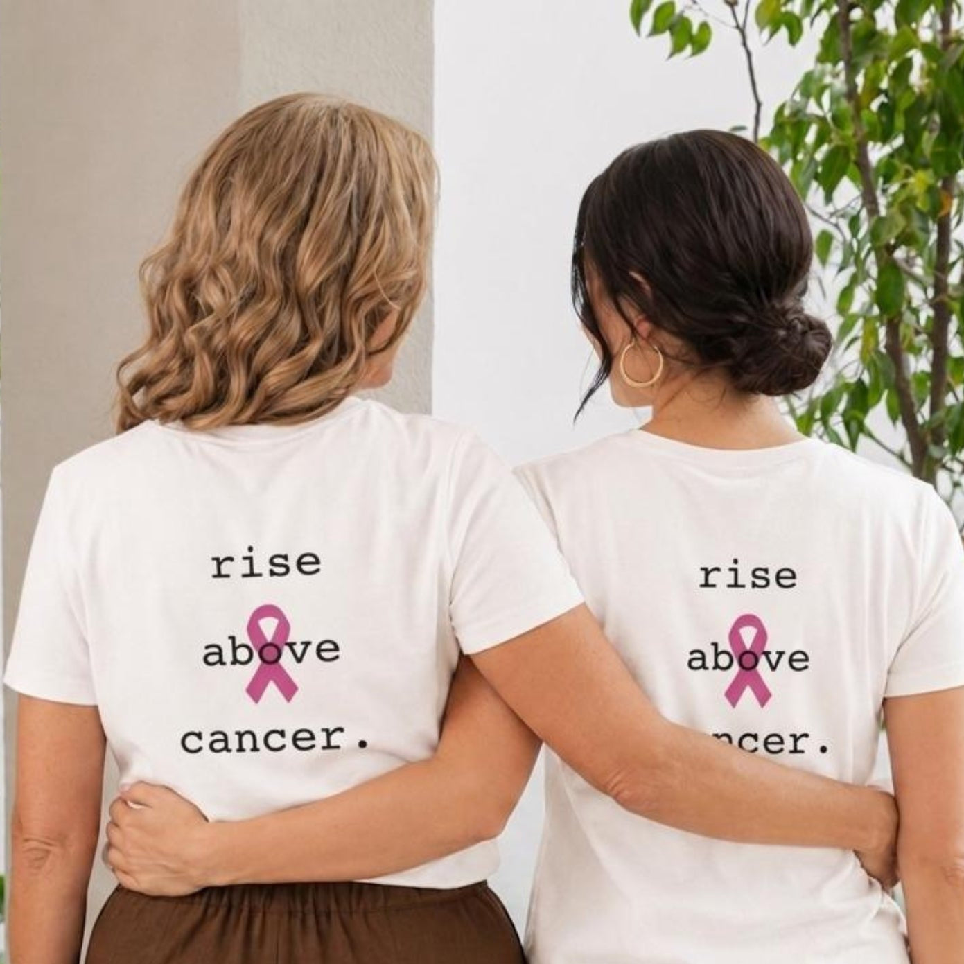 Two women standing side by side from the back wearing matching white graphic t-shirts with the text 'rise above cancer.' and a pink ribbon, paired with brown and beige pants, in an outdoor garden setting.