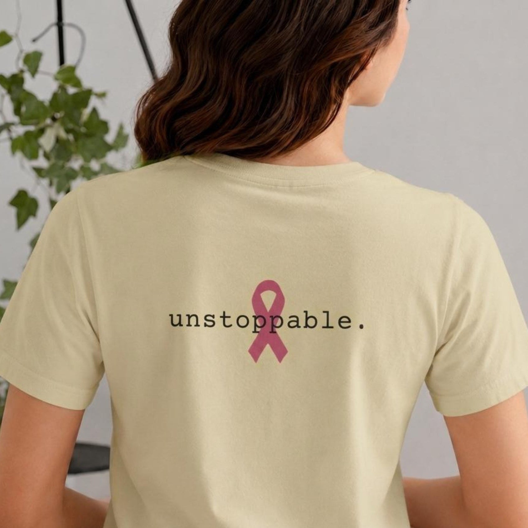 A woman viewed from behind wearing a light natural-colored short-sleeve T-shirt with the word 'unstoppable.' in black text and a pink awareness ribbon graphic printed on the back.