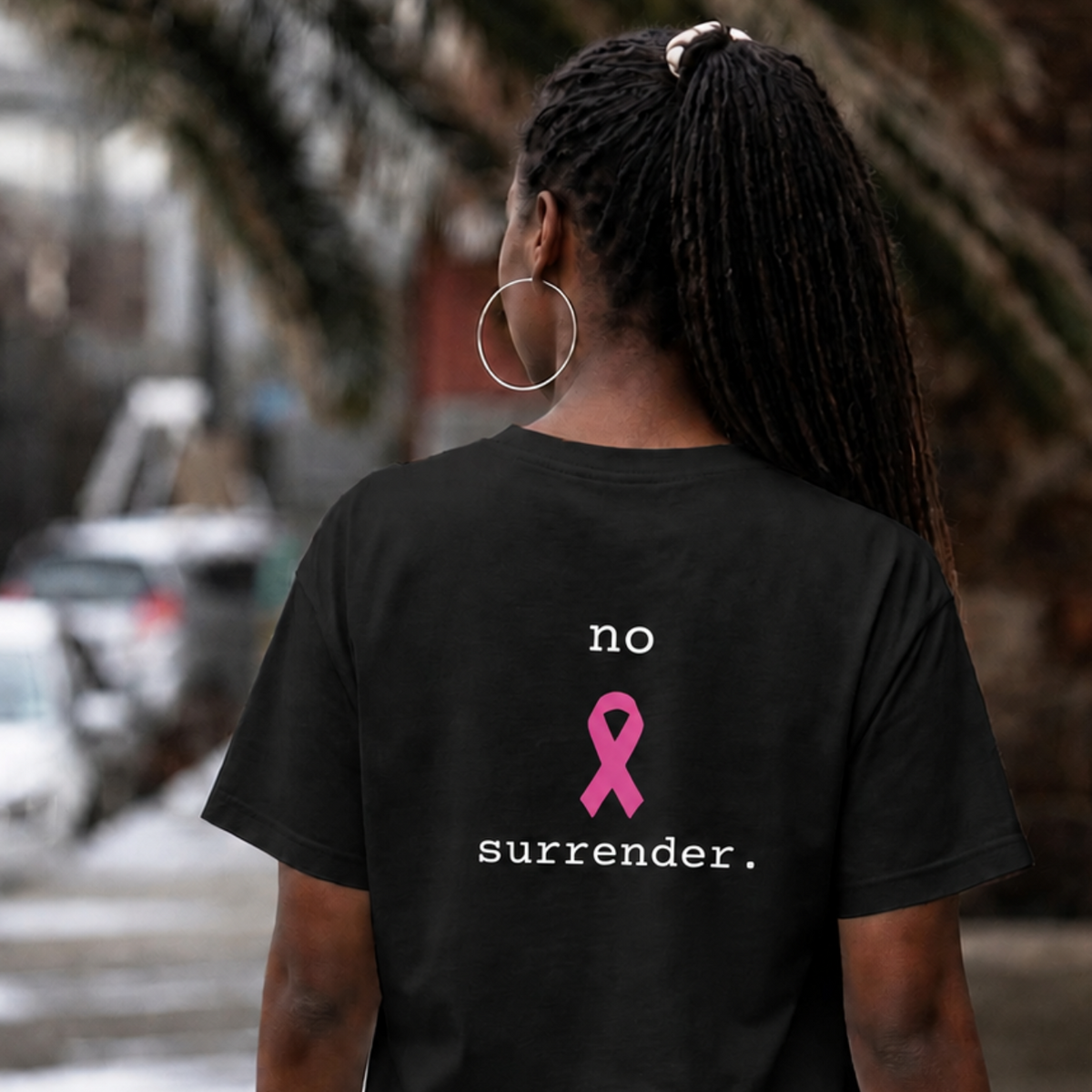 A woman viewed from behind wearing a black t-shirt with a pink ribbon graphic and the text 'no surrender.' printed on the back, set against a blurred outdoor street background.