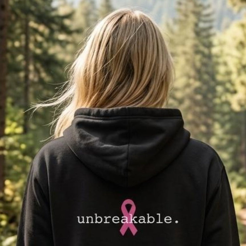 A woman viewed from behind stands on a forest path wearing a black hoodie with the word 'unbreakable.' in white and a pink ribbon graphic on the back, with mountains and trees in the background.