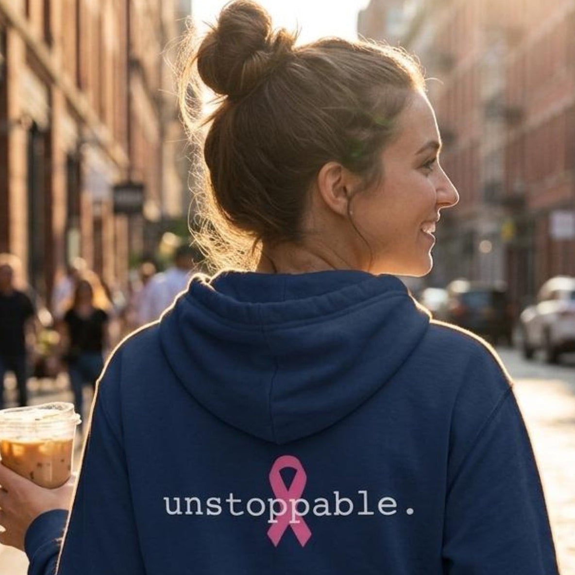 A woman on a city street wearing a classic navy blue hoodie with a pink ribbon and the text 'unstoppable.' on the back, holding an iced coffee.