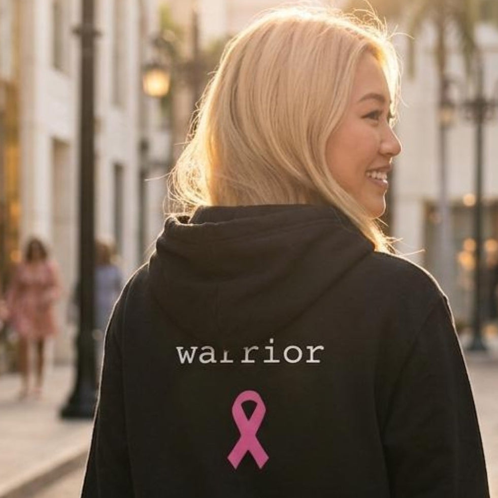 A woman viewed from behind wearing a black hoodie with 'warrior mode.' and a pink ribbon printed on the back, walking down a sunlit upscale street with luxury storefronts in the background.