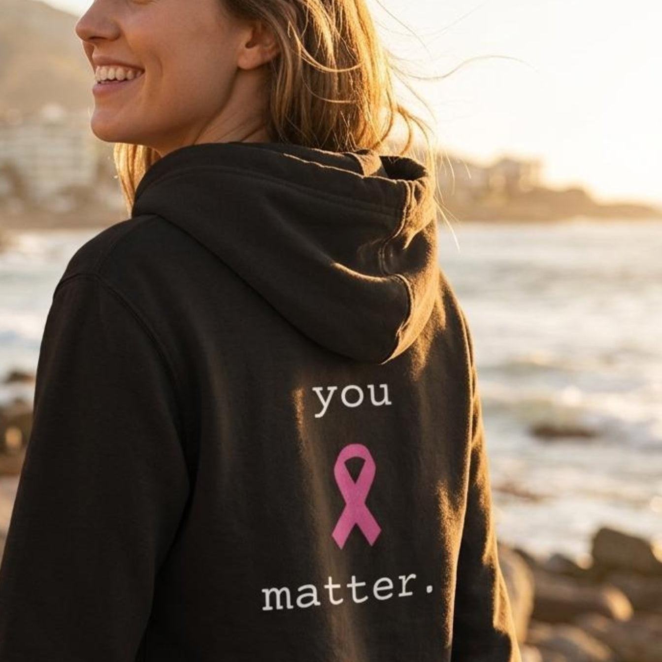 A woman wearing a black hoodie with a pink ribbon and the words 'you matter.' printed on the back, standing on a beach path with the ocean in the background.