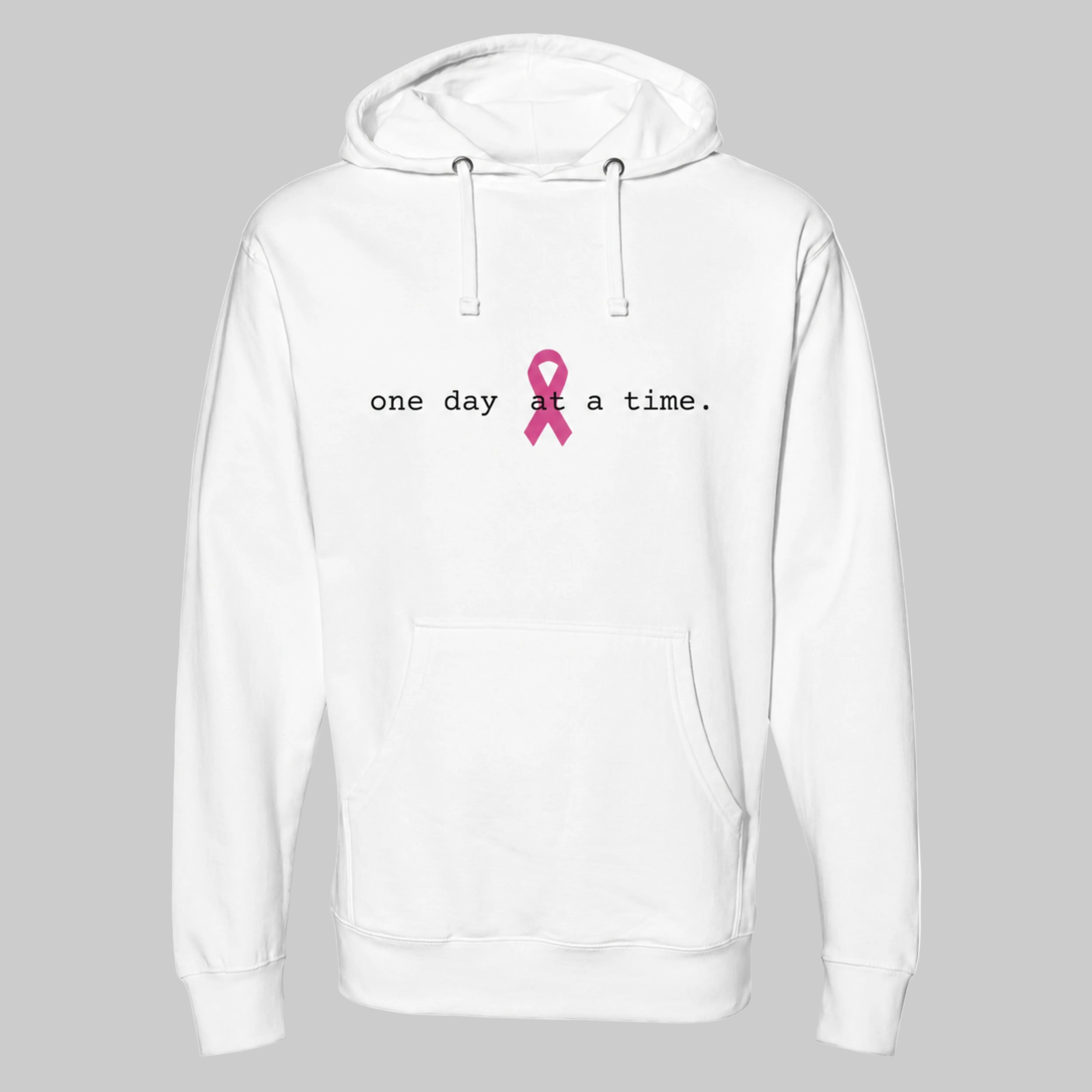 A white pullover hoodie with a front kangaroo pocket and drawstring hood, featuring the printed text 'one day at a time.' in black next to a pink awareness ribbon on the chest on a warm gray-colored background.