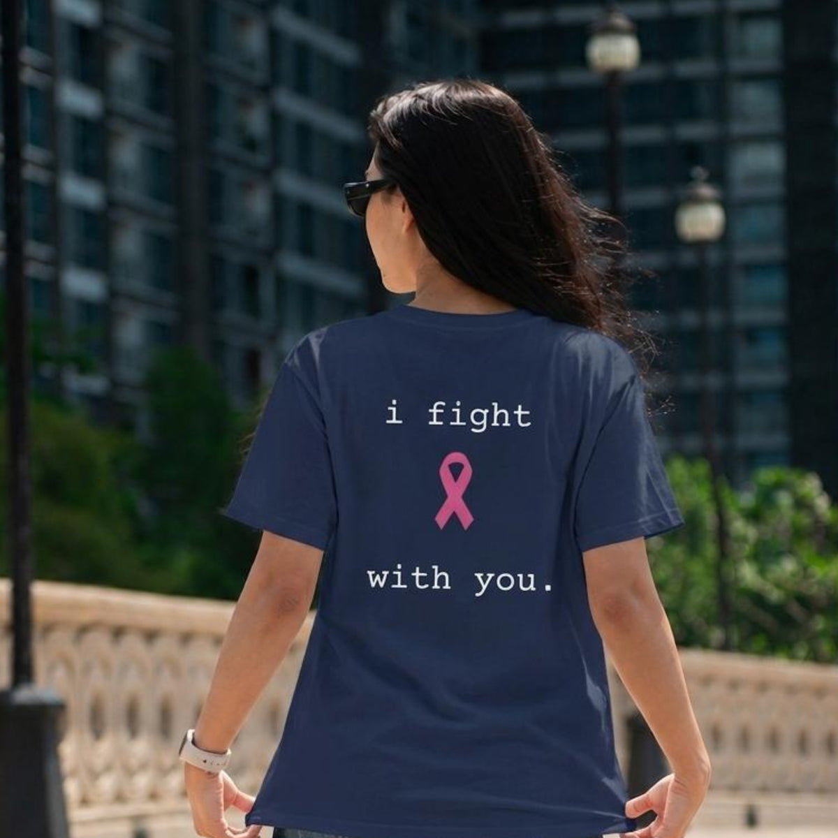 A woman viewed from behind wearing a navy blue t-shirt with a pink awareness ribbon and the text 'i fight with you.' printed on the back, set against a blurred urban background.
