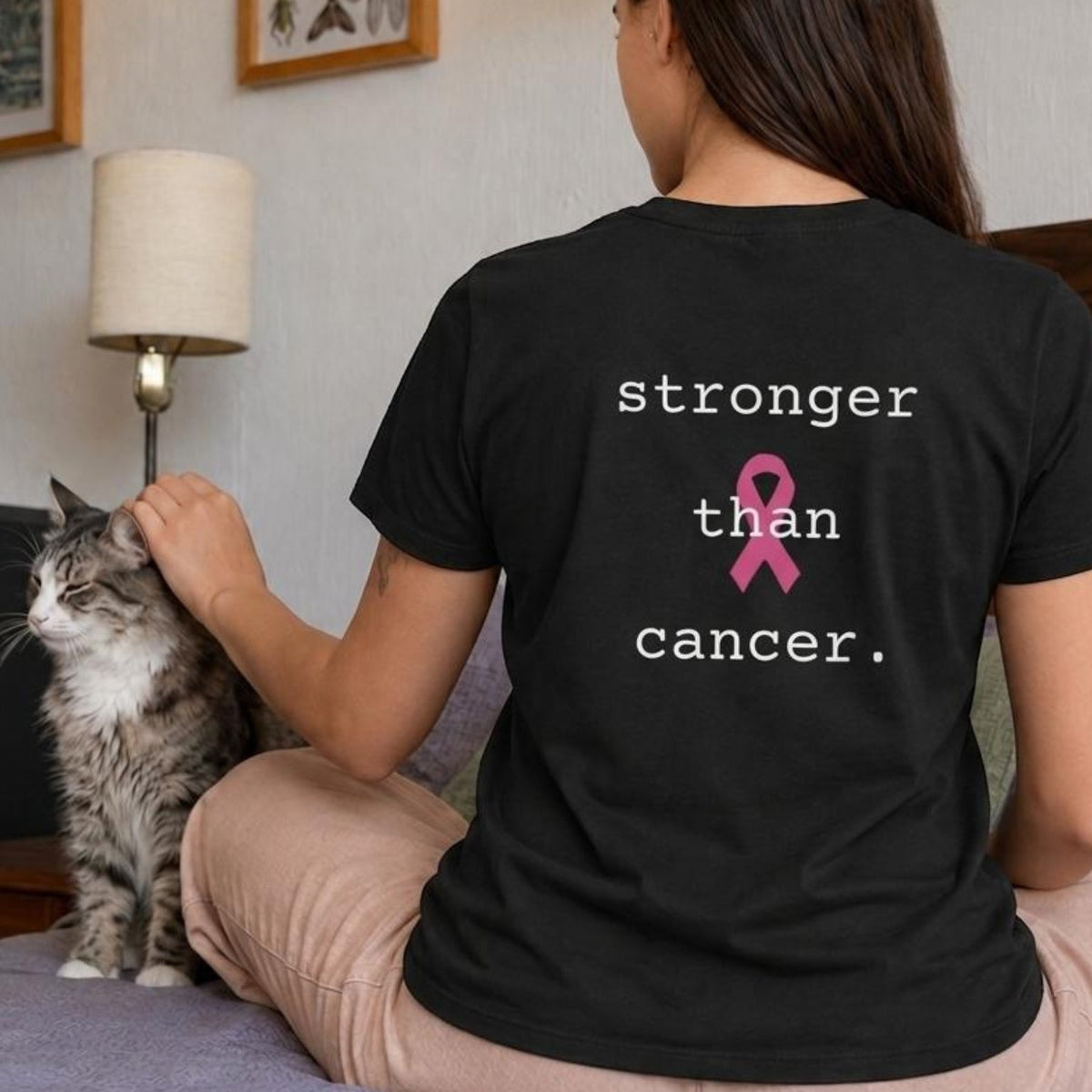 A woman sitting on a bed facing away from the camera, wearing a black t-shirt with the printed text 'stronger than cancer.' and a pink ribbon on the back.