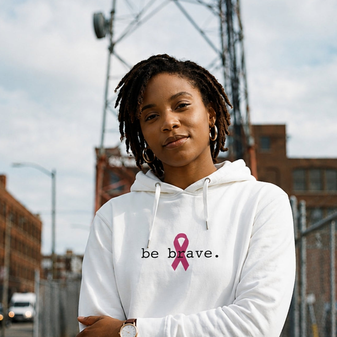 A woman with dreadlocks stands outdoors wearing a white hoodie with the text 'be brave.' and a pink ribbon printed on the chest.