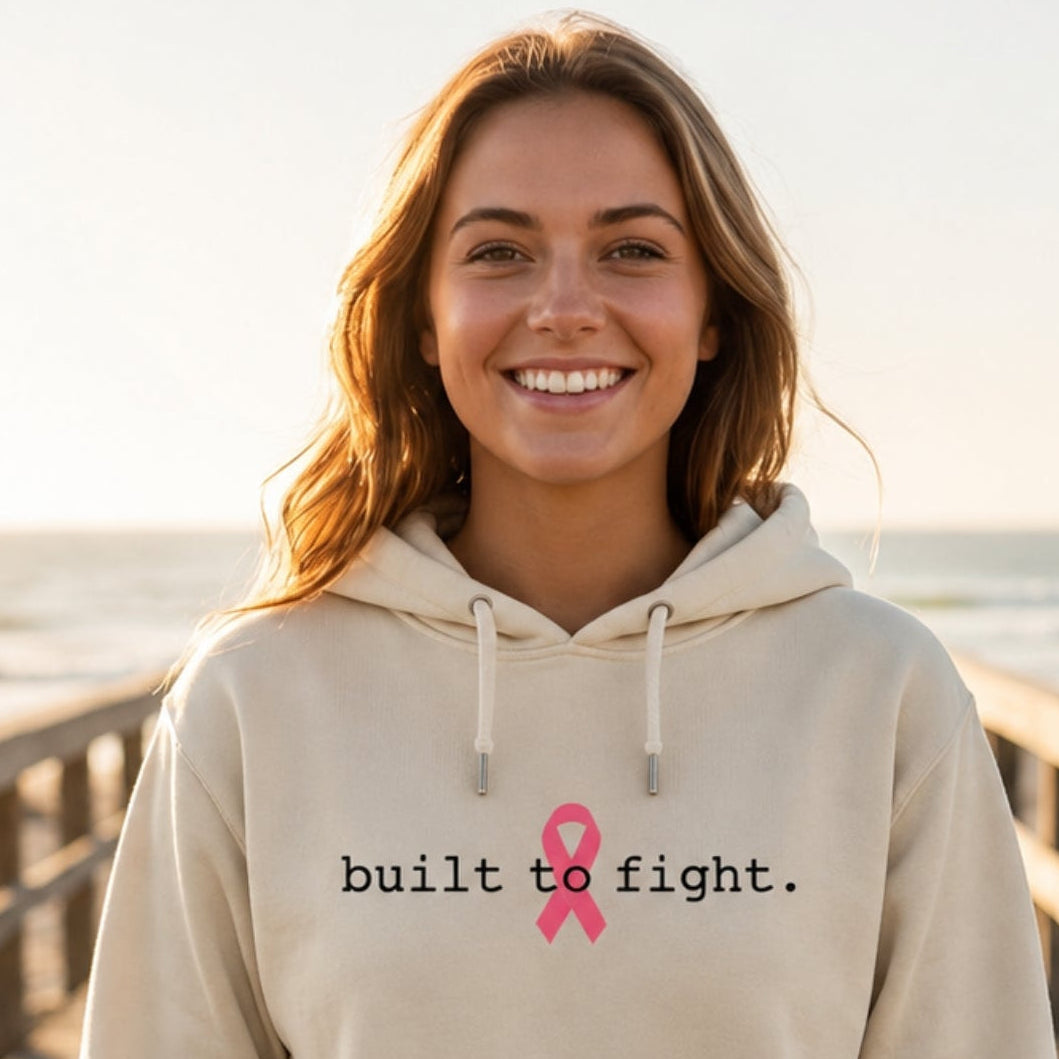 A smiling woman stands on a wooden boardwalk by the ocean wearing a bonr-colored hoodie with the text 'built to fight.' and a pink ribbon graphic on the chest.