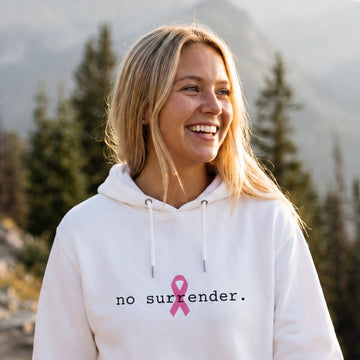 A woman smiling on a mountain trail wearing a white hoodie with the text 'no surrender.' and a pink ribbon graphic on the chest, with pine trees and mountains in the background.