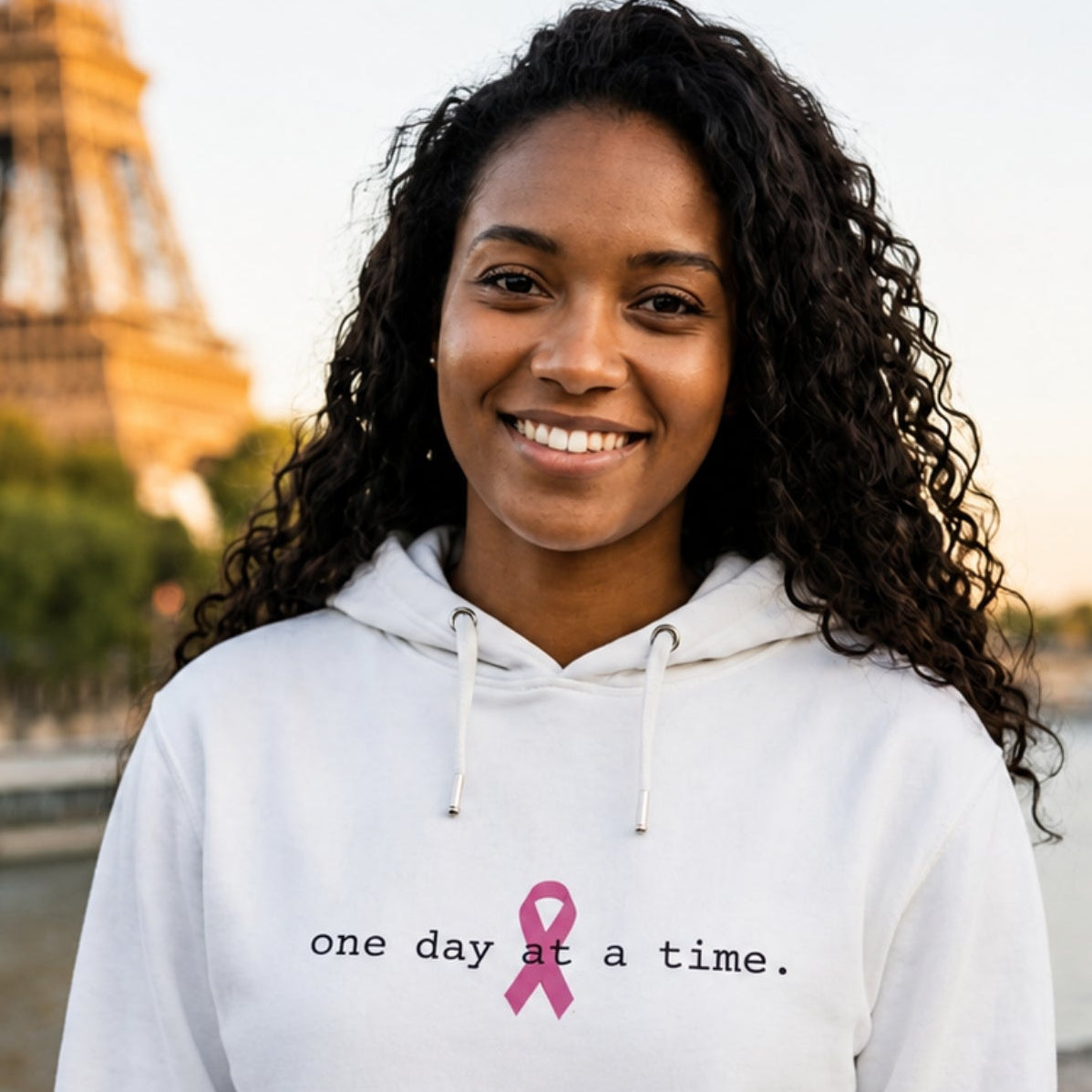 A smiling woman with curly dark hair stands outdoors in front of the Eiffel Tower, wearing a white hoodie with the text 'one day at a time.' and a pink ribbon printed on the chest.