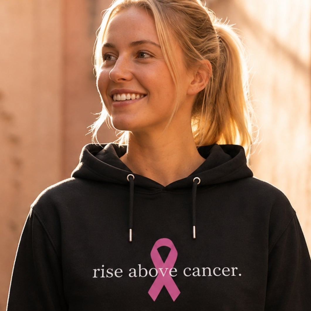 A woman wearing a black hoodie with a pink ribbon and the text 'rise above cancer.' printed on the front, standing in a sunlit outdoor alley with warm beige walls.