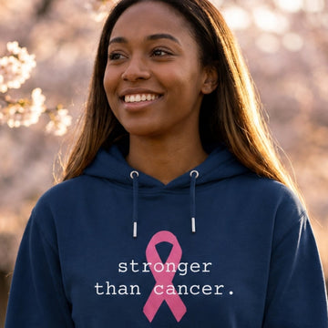 A woman stands on a path surrounded by blooming cherry blossom trees, wearing a navy blue hoodie with a pink ribbon graphic and the text 'stronger than cancer.' printed on the front.