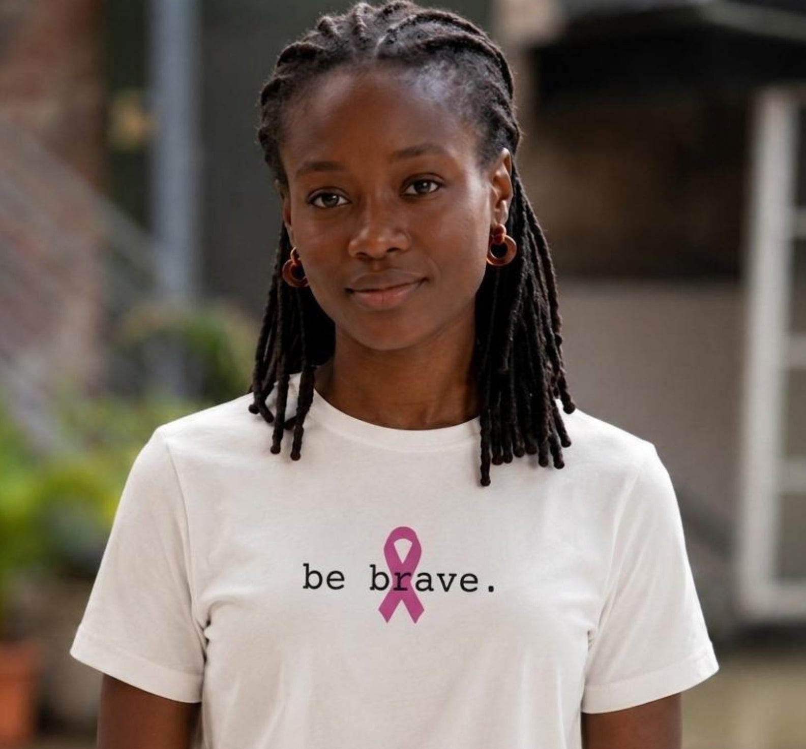 A woman with black dreadlocks stands wearing a white short-sleeve t-shirt with the text 'be brave.' and a pink ribbon printed on the chest.