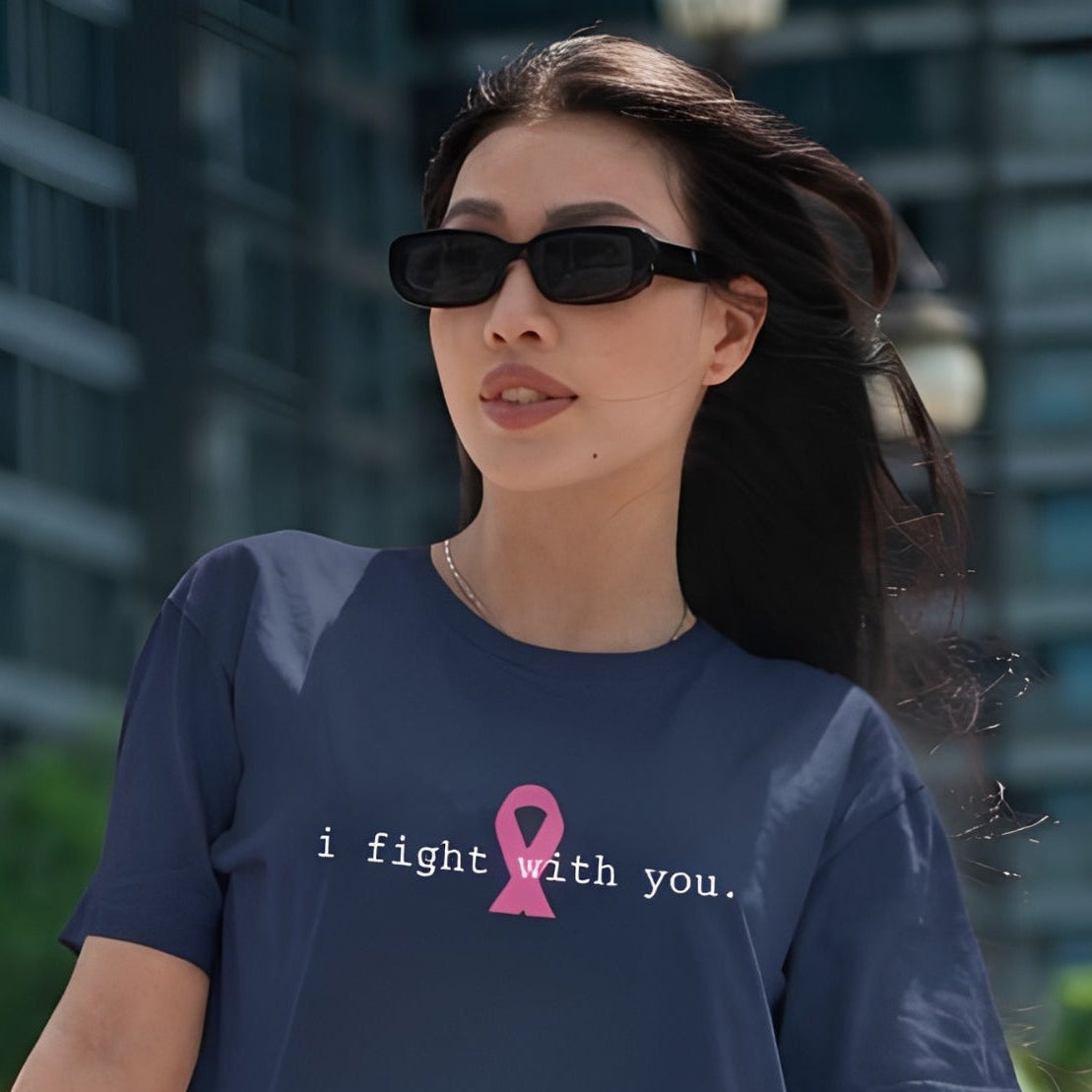 A woman wearing a navy blue graphic t-shirt with a pink ribbon and the text 'i fight with you.' printed on the chest, set against an outdoor urban background with buildings and trees.