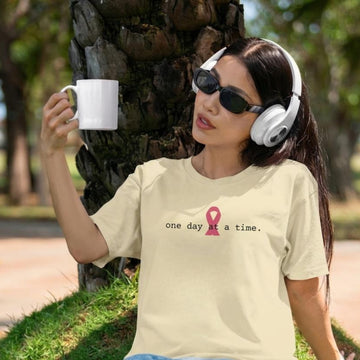 A woman outdoors wearing a natural t-shirt with a pink awareness ribbon and the text 'one day at a time.' She is also wearing white over-ear headphones and holding a white mug near a tree trunk.