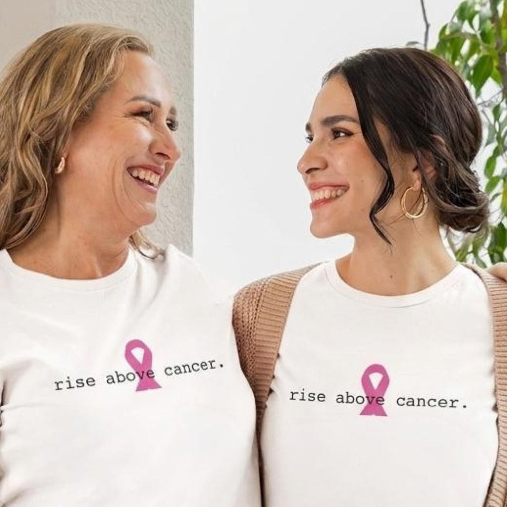Two women standing side by side wearing matching white graphic T-shirts with a pink ribbon and the text 'rise above cancer.' They are posed outdoors with plants and a gift box in the background.