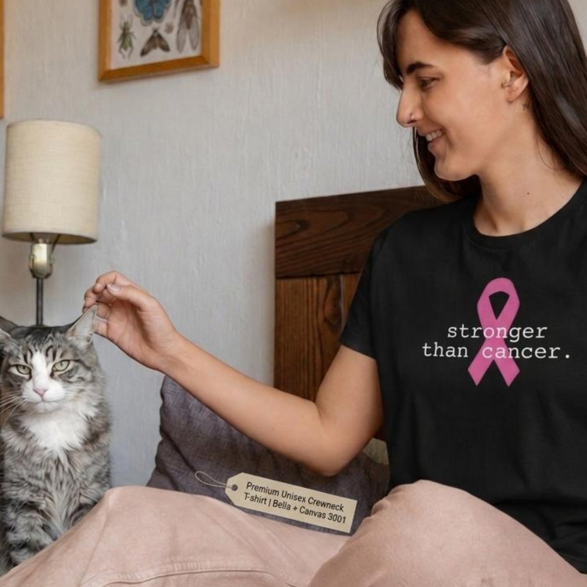 A woman sitting on a bed in a bedroom, wearing a black t-shirt with a pink ribbon and the text 'stronger than cancer.' She is petting a gray tabby cat.
