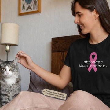 A woman sitting on a bed in a bedroom, wearing a black t-shirt with a pink ribbon and the text 'stronger than cancer.' She is petting a gray tabby cat.