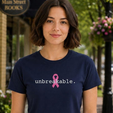 A woman wearing a navy blue t-shirt with a pink ribbon graphic and the word 'unbreakable.' printed in white, standing on a city sidewalk in front of a storefront.
