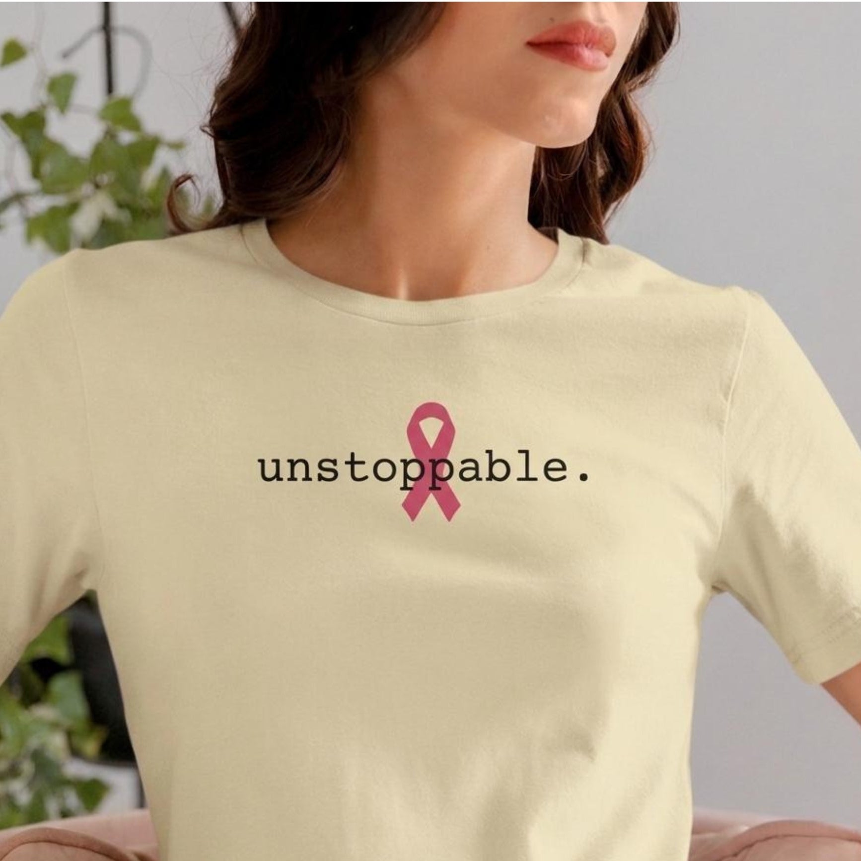 A woman wearing a natural-colored t-shirt with a pink ribbon graphic and the word 'unstoppable.' printed in black on the chest, set against a soft-focus indoor background.