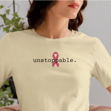 A woman wearing a natural-colored t-shirt with a pink ribbon graphic and the word 'unstoppable.' printed in black on the chest, set against a soft-focus indoor background.