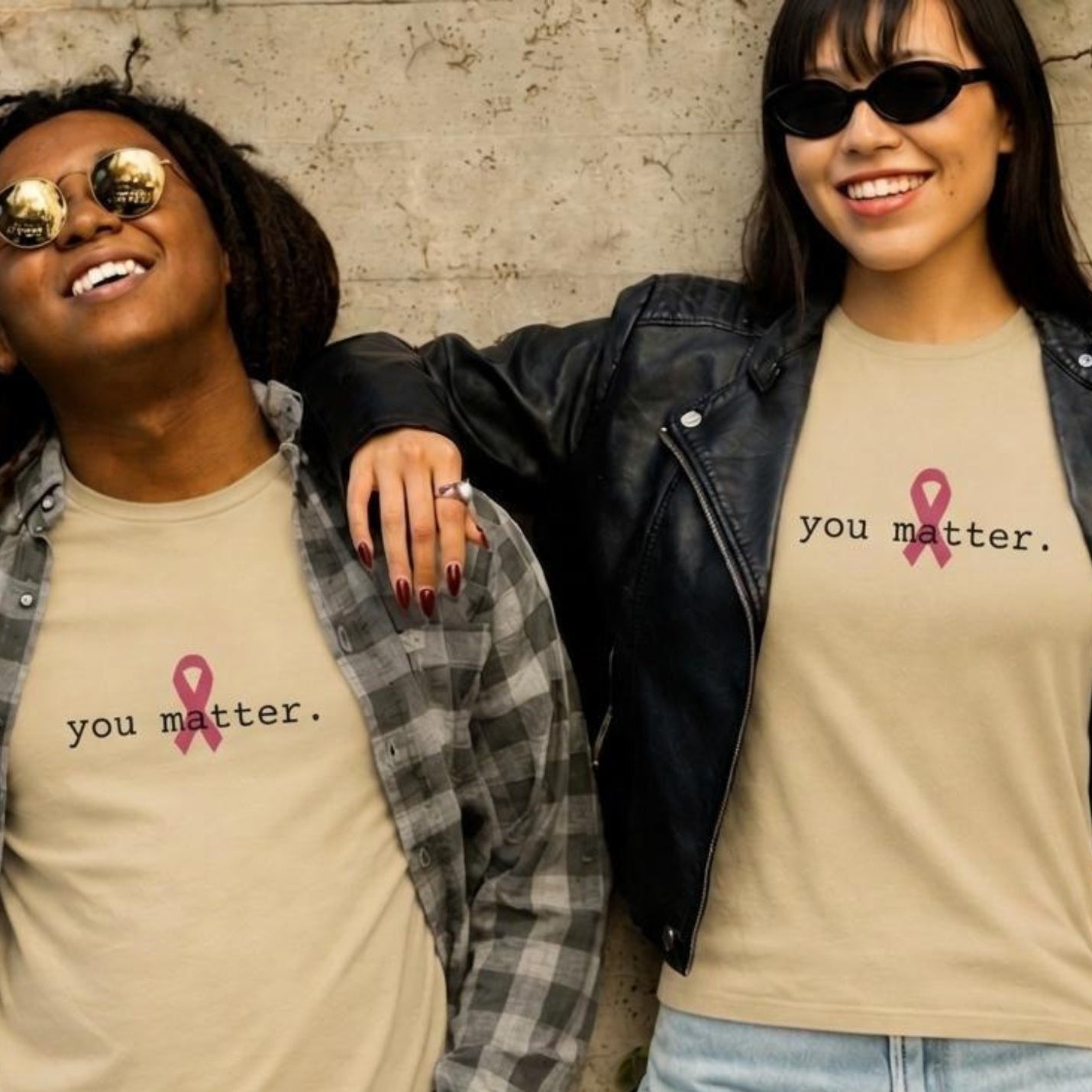 Two women standing together against a textured concrete wall, both wearing natural-colored graphic t-shirts with the text 'you matter.' and a pink ribbon, styled with different outerwear (a plaid shirt and a black leather jacket).