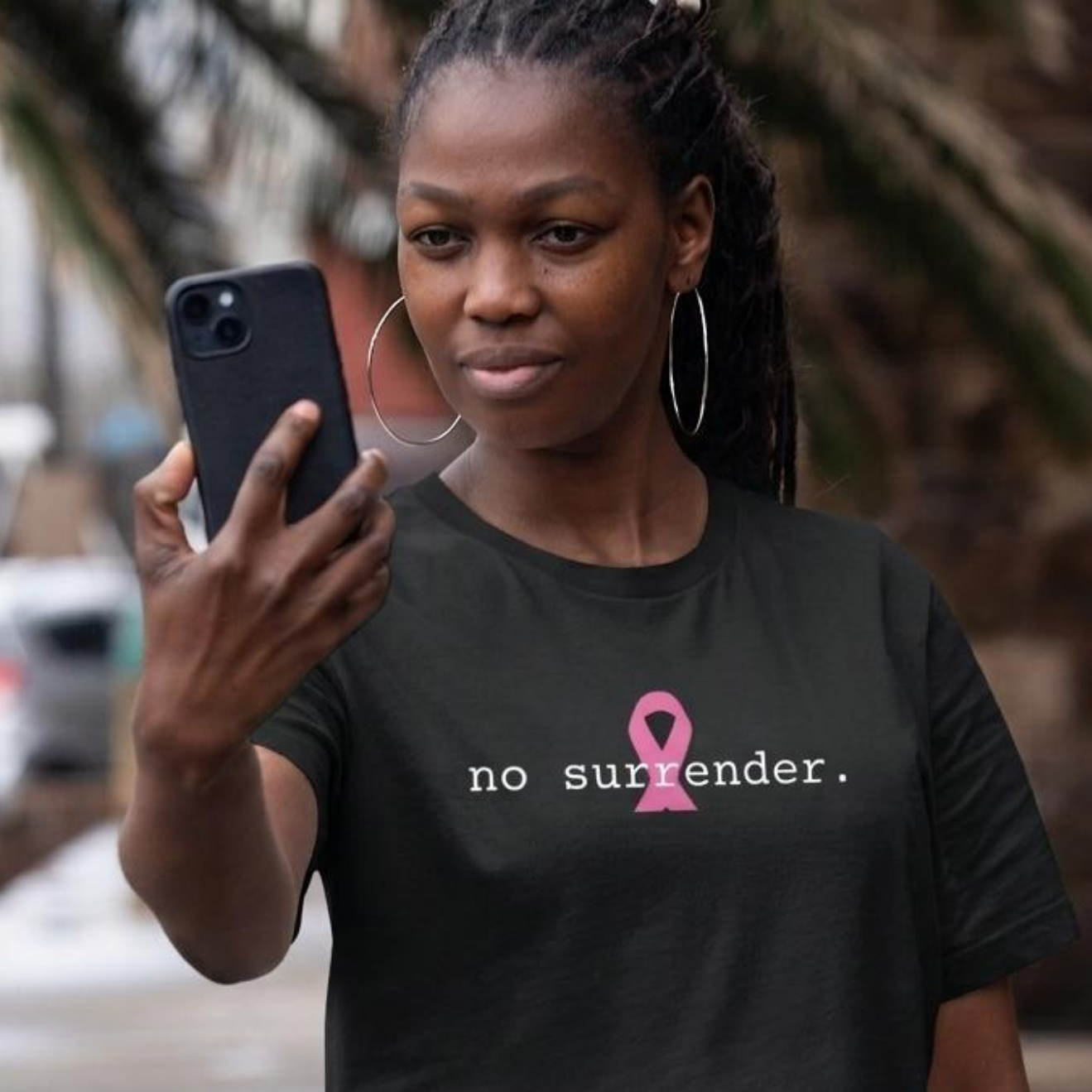 A woman taking a mirror selfie outdoors wearing a black t-shirt with a pink ribbon graphic and the text 'no surrender.' printed on the chest.