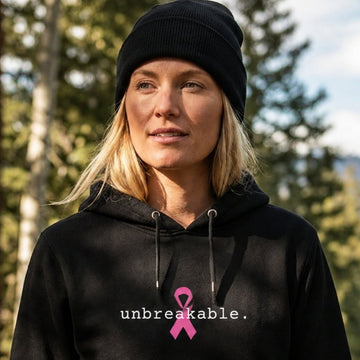 A woman wearing a black beanie and black hoodie stands outdoors on a forest path. The hoodie has a pink ribbon graphic and the word 'unbreakable.'