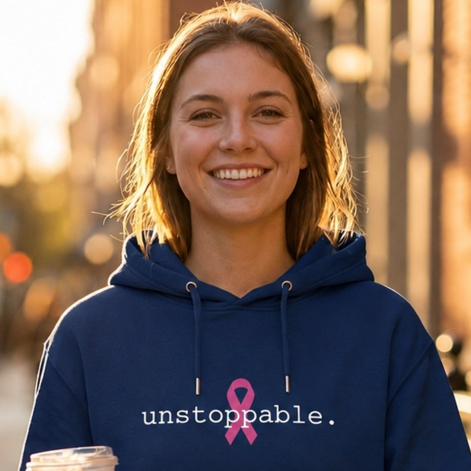 A woman walking on a sunlit city sidewalk wearing a classic navy blue hoodie with a pink ribbon and the word 'unstoppable.' printed on the chest, while holding an iced coffee.