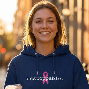 A woman walking on a sunlit city sidewalk wearing a classic navy blue hoodie with a pink ribbon and the word 'unstoppable.' printed on the chest, while holding an iced coffee.