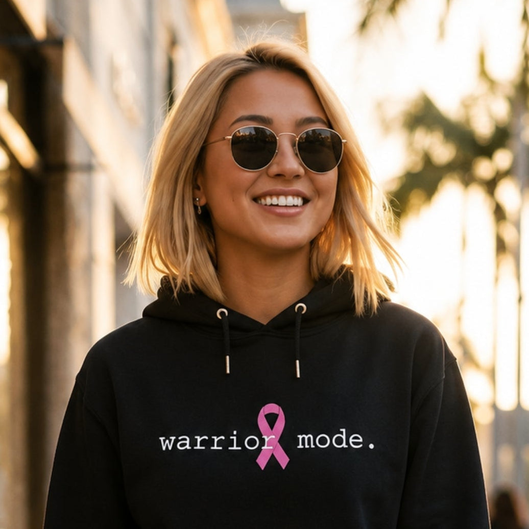 A woman wearing a black hoodie with the text 'warrior mode.' and a pink ribbon graphic walks down a sunlit city sidewalk lined with palm trees.
