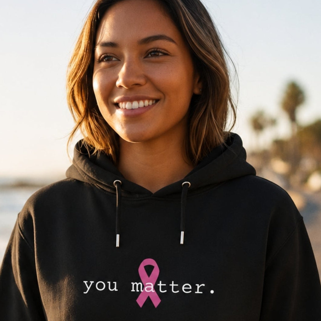 A woman wearing a black hoodie with the text 'you matter.' and a pink awareness ribbon printed on the chest, standing on a coastal road with the ocean in the background.
